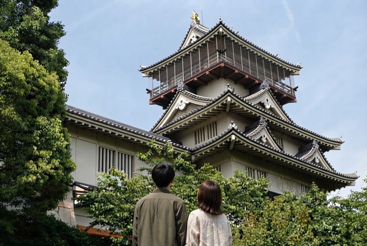 緑の木々の向こうにそびえる岩崎城の展望塔を見上げるカップルの後ろ姿